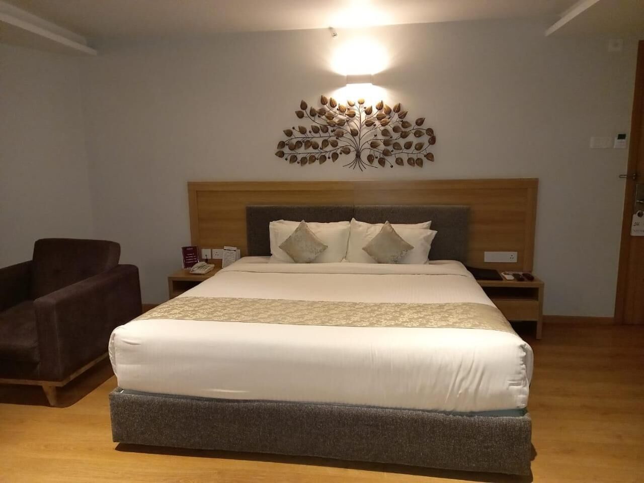 A hotel room with a large bed, a wooden headboard, a wall-mounted light fixture, a brown armchair, and a bedside table with a phone.