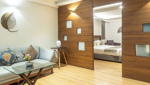 A modern hotel room with a wooden partition, a glass coffee table, a sofa with decorative pillows, and a bed with a grey headboard. - Native By Chancery Hotels (Belagavi)