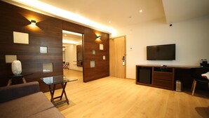 A modern living room with a wooden floor, a glass coffee table, a sofa, a TV, and a wooden cabinet. - Native By Chancery Hotels (Belagavi)
