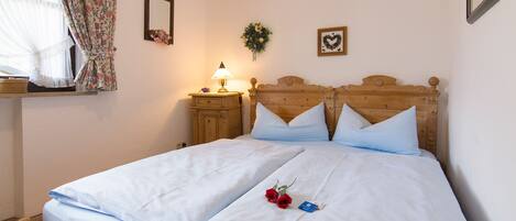 Una cama bien hecha con sábanas blancas y dos almohadas, una mesita de noche con lámpara, y una cortina con estampado floral.
