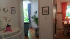 A room with a white cabinet, a small table with a pink pot, a patterned rug, and a doorway leading to another room with a dining table and chairs.