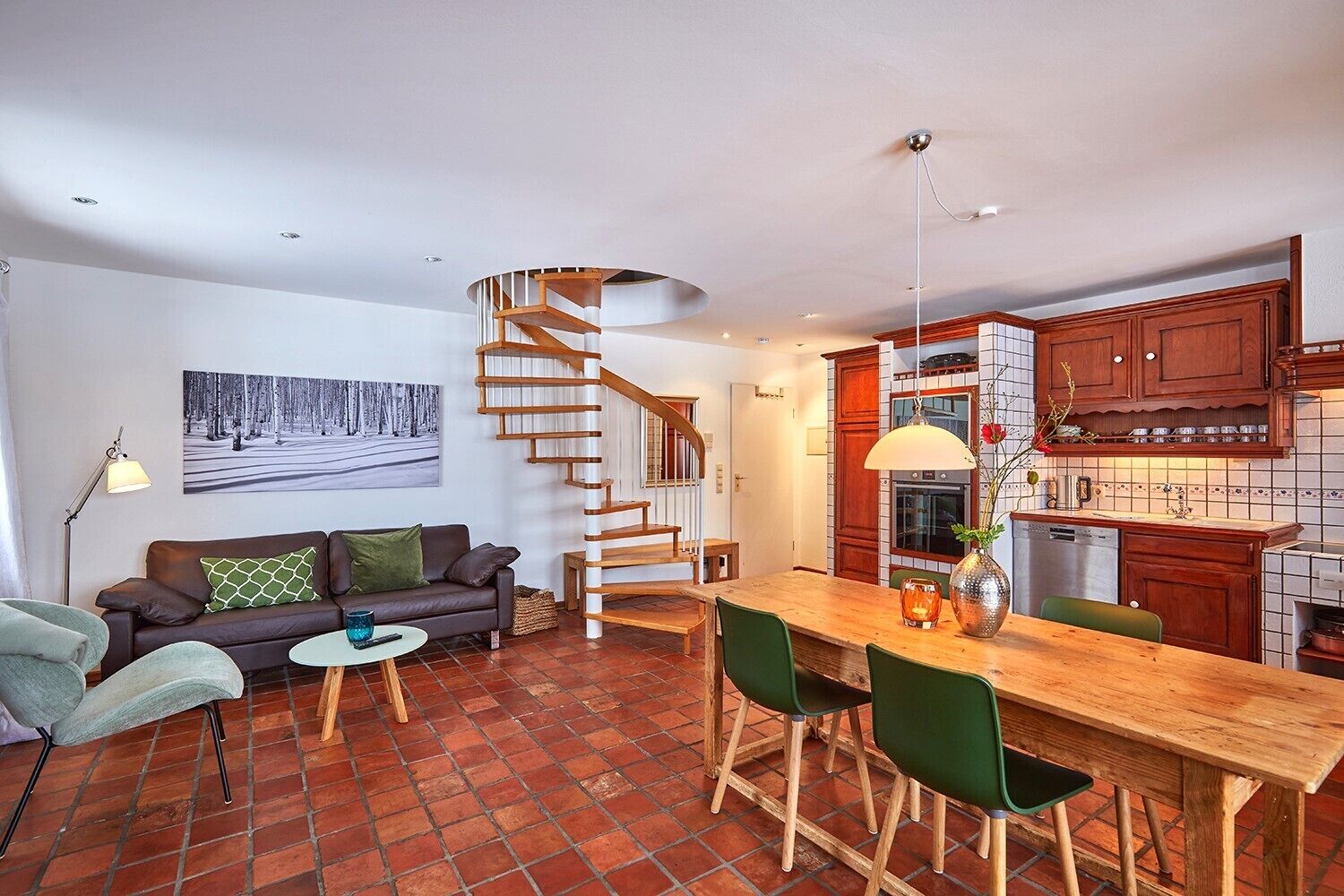 A modern living room with a wooden staircase, a dining area with a wooden table and green chairs, and a kitchen with wooden cabinets and a tiled backsplash.