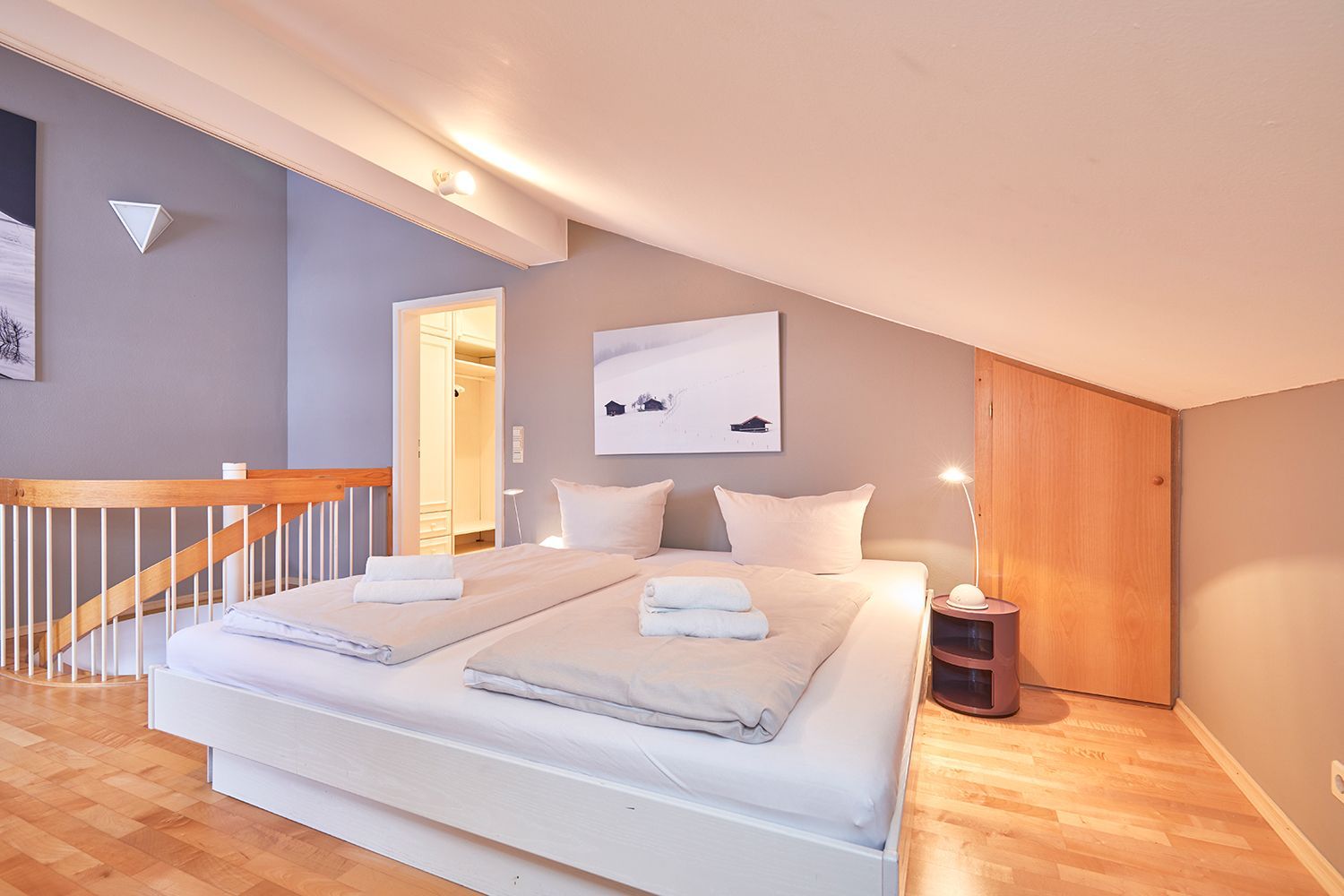 A double bed with white bedding and two pillows, a bedside table with a lamp, a wooden floor, a wooden railing, and a door leading to a bathroom.