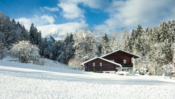 Eine Holzhütte mit geneigtem Dach, umgeben von schneebedeckten Bäumen und wolkenlosem Himmel.