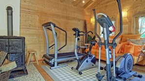 Ein Holzblockhaus-Innenraum mit Fitnessbereich, der Laufbänder und ein Crosstrainer zeigt, einem gemütlichen Wohnbereich mit einer Couch und Kissen sowie einem Kamin mit Holzscheiten.