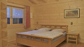 Camera da letto in una baita di legno, con un letto, comodini, una finestra con tende e un quadro appeso al muro.