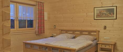A wooden cabin bedroom with a bed, bedside tables, a window with curtains, and a painting on the wall.