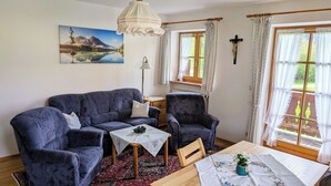 A living room with a sofa, armchairs, a small table, and a dining table with a vase. There is a painting on the wall and a cross above the dining table. - Apartment 2, 55sqm, balcony, kitchenette, 2 bedrooms, 1 living room, WLAN (Ruhpolding)
