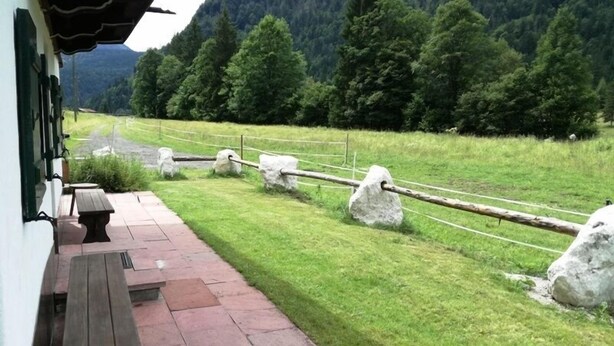 Un patio con un banco de madera y una mesa, un muro de piedra con cerca de cuerda, y un campo cubierto de césped con árboles y montañas al fondo.