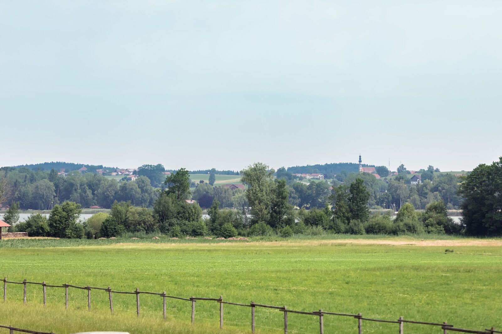 Paisagem rural com cerca, árvores e uma cidade distante com edifícios e uma igreja.