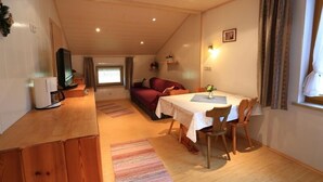 A cozy room with a dining table, chairs, a sofa, a TV, and a window with curtains. - Zwei-Raum-Ferienwohnung "Kaiserblick" 46qm, Dusche/WC, Extra-Schlafraum, Küche (Reit im Winkl)