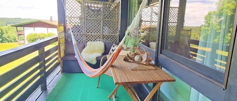 A balcony with a hammock, a wooden table, and a view of the outdoors.