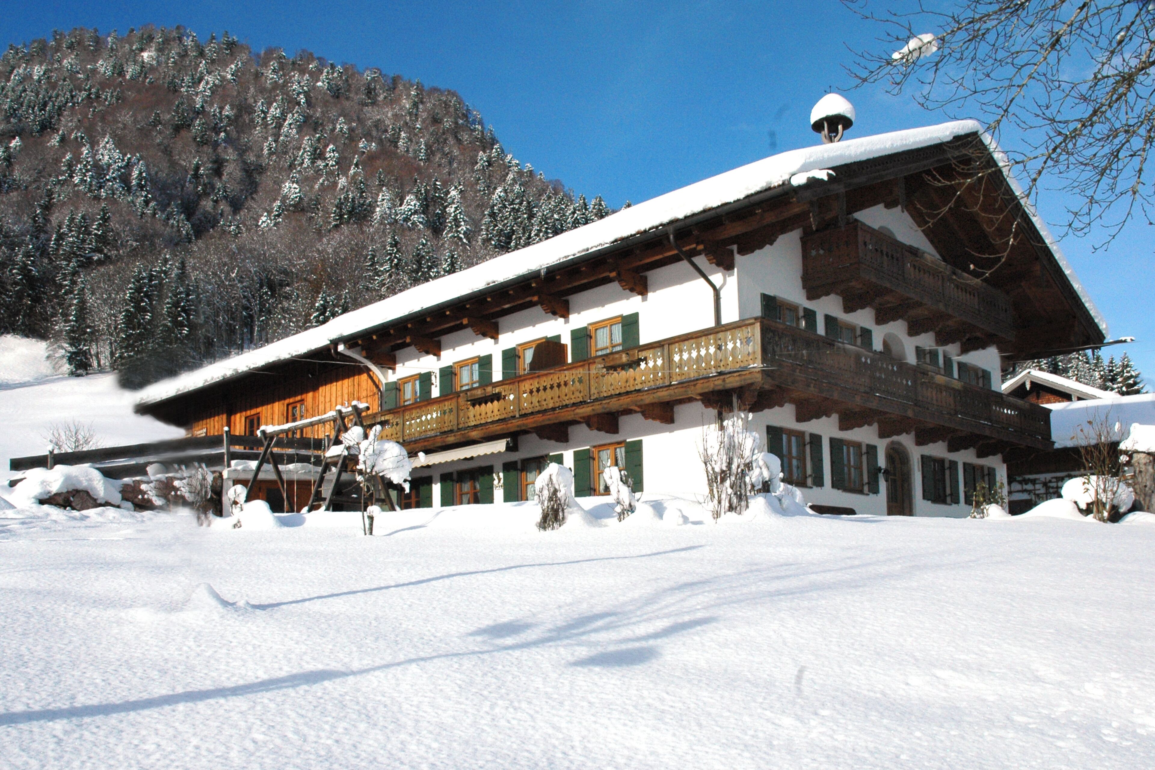 Un chalet traditionnel doté d’un balcon en bois, entouré d’arbres couverts de neige et avec une chaîne de montagnes en toile de fond.