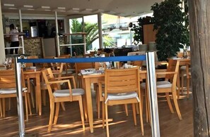 A restaurant with wooden tables and chairs, a kitchen area, and large windows.