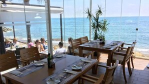 A restaurant with large windows overlooking the ocean, wooden tables and chairs, and a tropical plant.