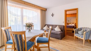 A cozy living room with a wooden floor, a dining table with chairs, a sofa, and a bed in the background. - Hausberg two-room vacation apartment 45 sqm (Reit im Winkl)