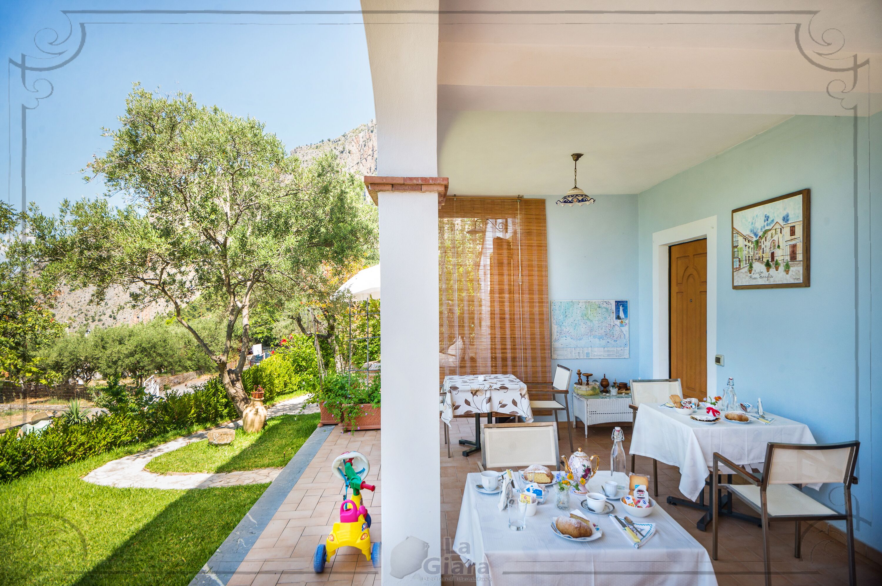 A dining area with tables set for a meal, a view of a garden with a pathway, and a child’s toy car.