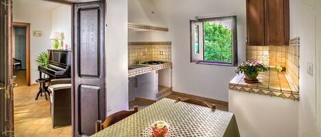 A compact kitchen with a dining area, a piano, and a window with a view of greenery.