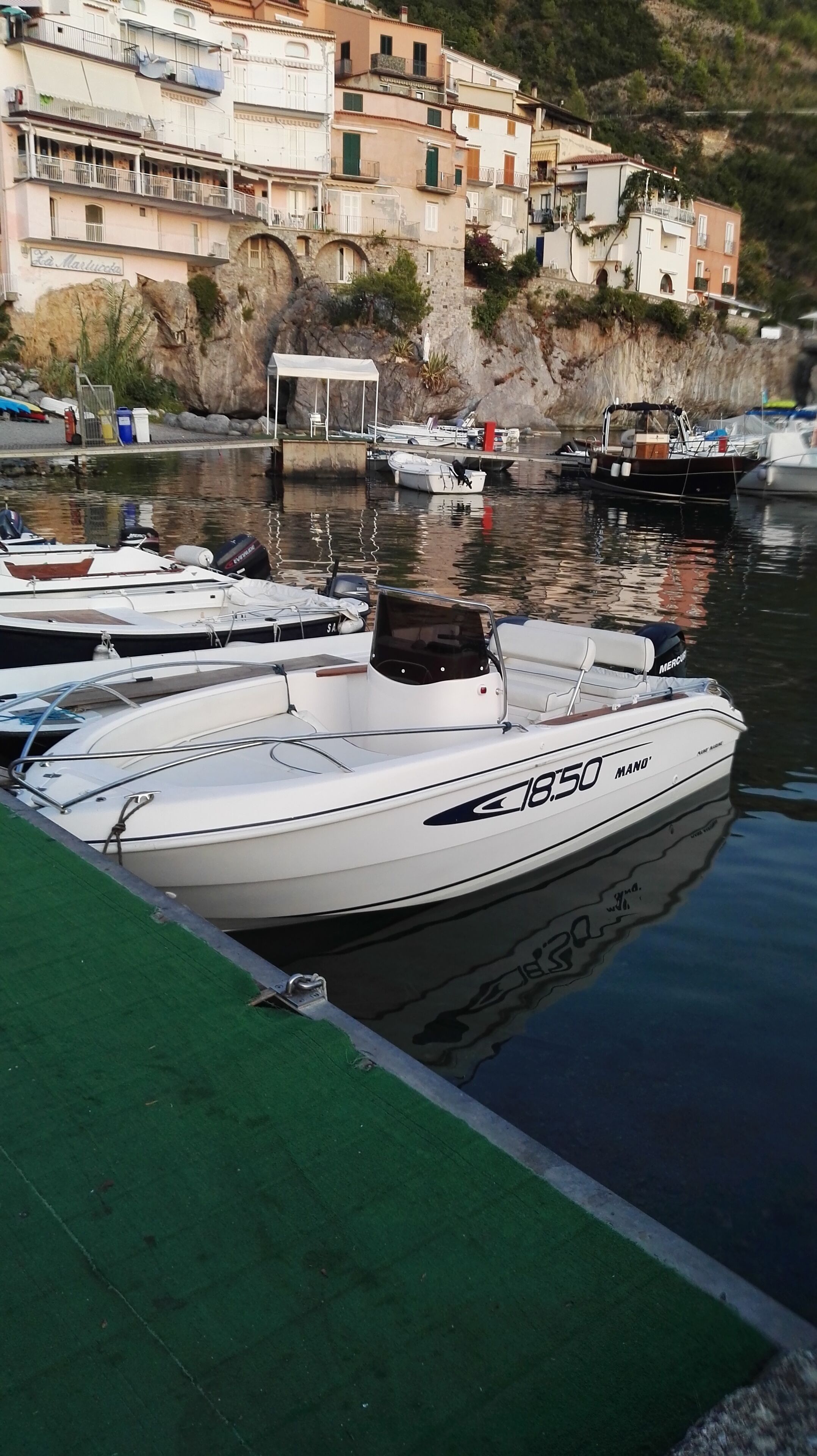 Un puerto deportivo con varios barcos amarrados, incluyendo un barco a motor blanco identificado con la leyenda "18.50 MANO".