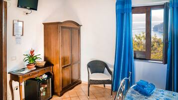 Una habitación con cama, una silla, escritorio con computadora, armario, ventana con cortinas y televisión en la pared.
