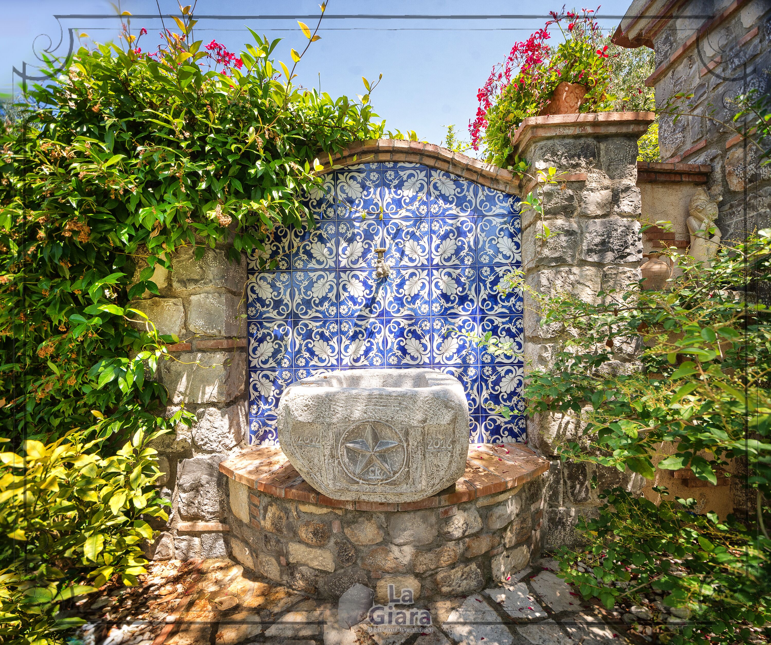 A stone wall with a blue and white tiled section, a stone bench, and surrounding greenery.