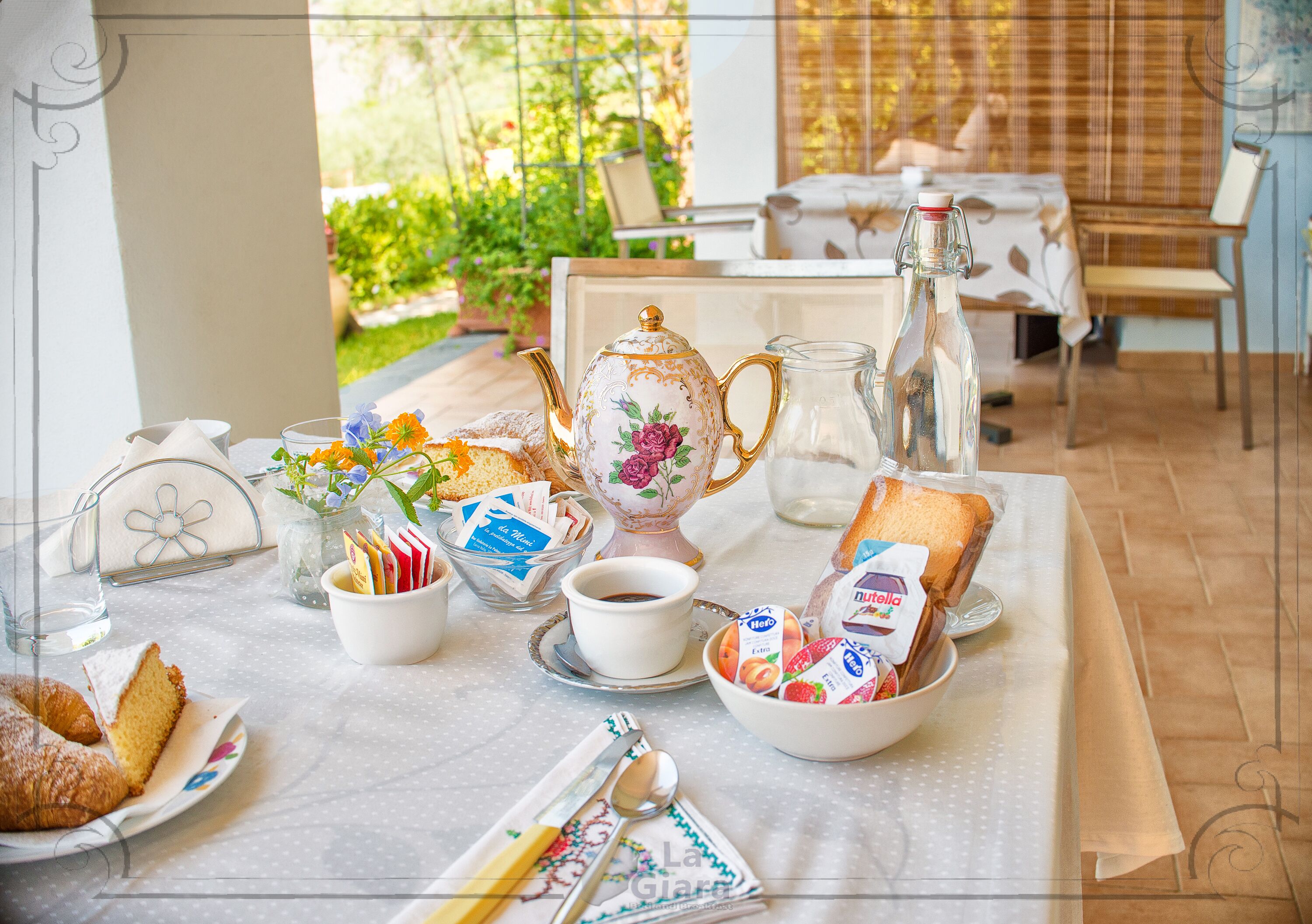 A breakfast table set with pastries, bread, and beverages, with a garden view in the background.