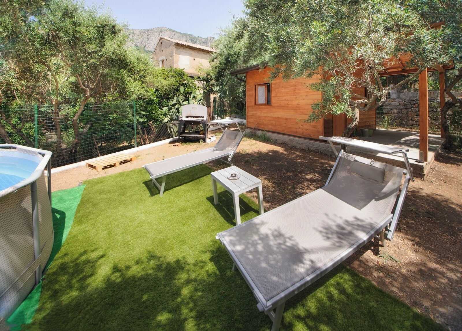 Un patio con césped artificial, dos sillones de descanso, una pérgola de madera y un pequeño edificio de madera.