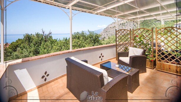 A covered outdoor seating area with wicker furniture, a glass table, and a view of the sea and hills. - B&B La Giara (Maratea)