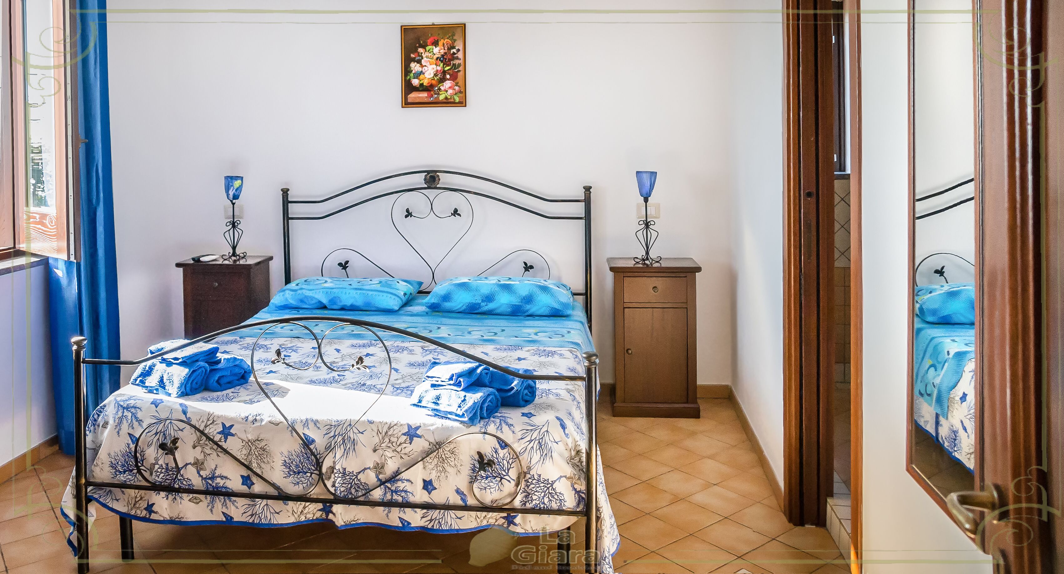Un lit bien fait, avec une literie bleue, une table de chevet en bois surmontée d’une lampe, et une petite armoire.