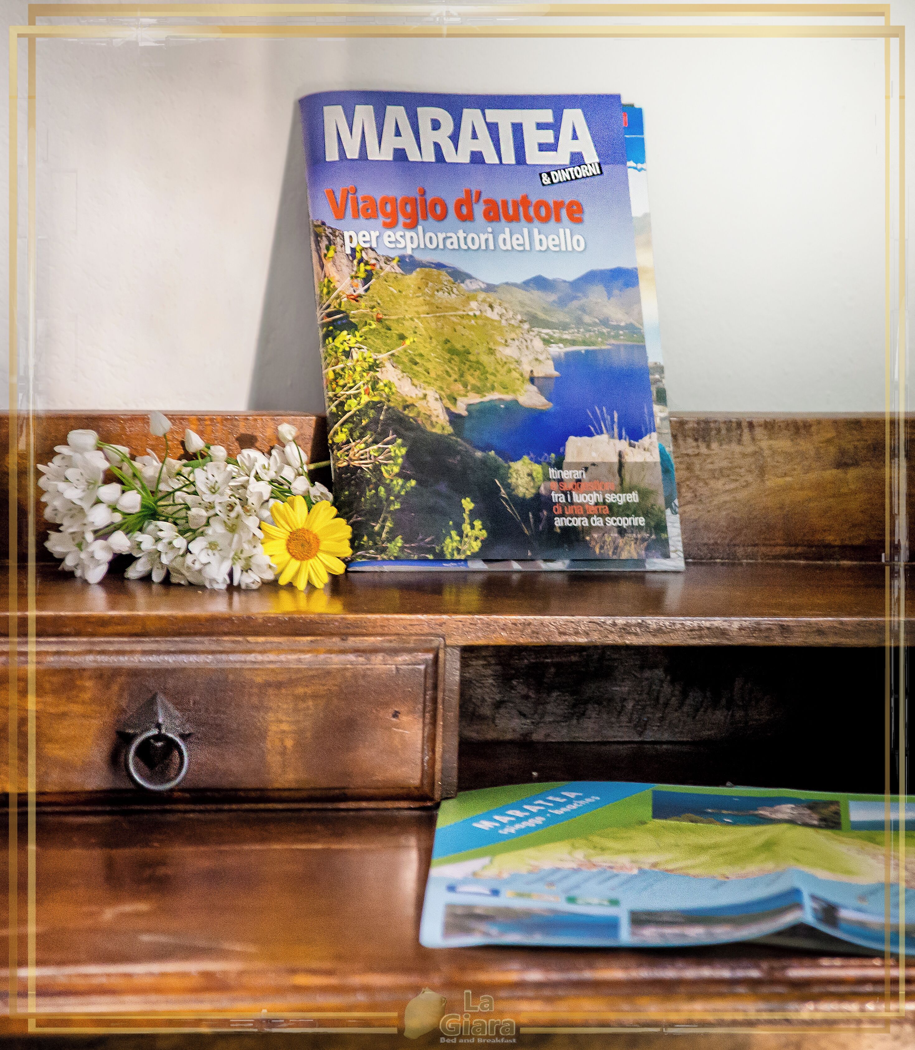Un libro titulado 'MARATEA & DINTORNI' sobre una superficie de madera, con un pequeño ramo de flores a su lado.