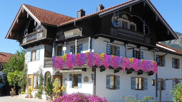 En traditionell byggnad i alphyttestil med en balkong som är prydd med färgstarka blommor, en skylt med texten ”HAUS EDEWEISS” och en klarblå himmel i bakgrunden.