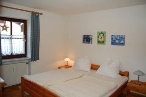 Ein Schlafzimmer mit einem Holzbett, weißer Bettwäsche, einem Nachttisch mit Lampe, einem Fenster mit Vorhängen und drei kleinen Wandbildern.