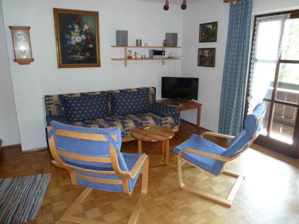 Un salon avec un canapé à motifs, deux fauteuils bleus, une table basse en bois, un téléviseur et une horloge au mur.