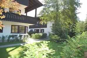 Ein zweistöckiges Gebäude mit Balkon, umgeben von einem gepflegten Garten und Bäumen.