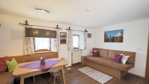 Ein gemütliches Wohnzimmer mit einem Holztisch, einer Couch und einem Sofa. An den Fenstern hängen Vorhänge, ein Heizkörper steht dort, und ein Bild ist an der Wand angebracht.
