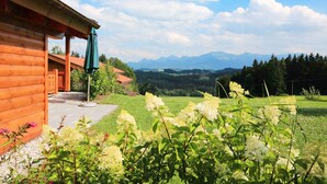A wooden cabin with a porch, surrounded by a garden with white flowers and a view of distant mountains. - Holzblockhaus Hochries ***** (Rimsting)
