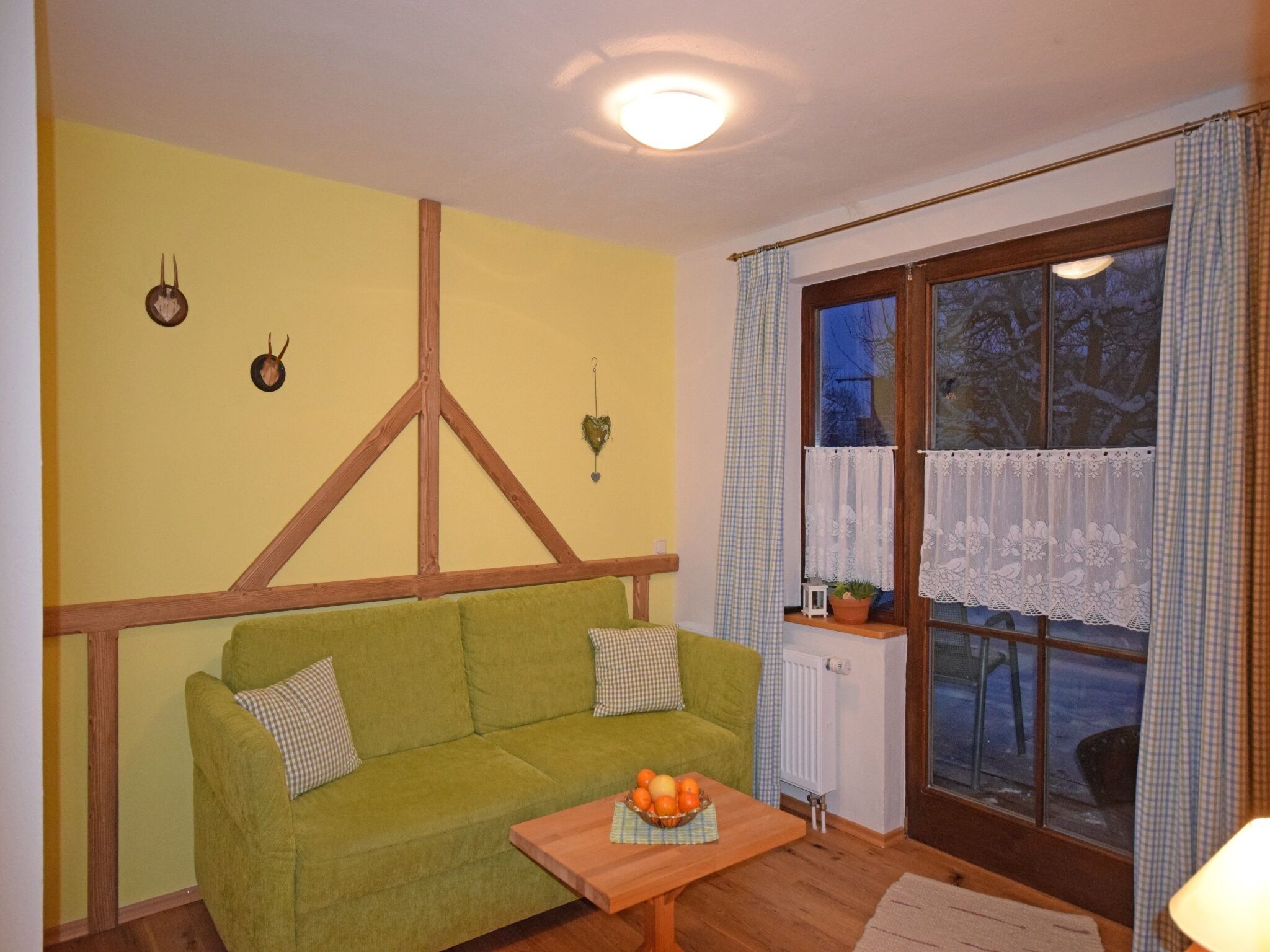 Una habitación acogedora con un sofá verde, una mesa de madera con fruta, una ventana con cortinas de encaje y un radiador.