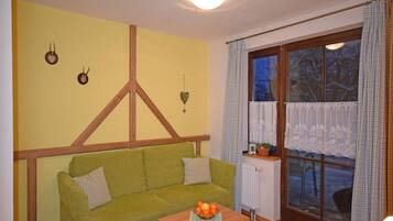 Una habitación acogedora con un sofá verde, una mesa de madera con fruta, una ventana con cortinas de encaje y un radiador.