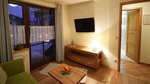 Ein gemütliches Wohnzimmer mit einem Holztisch, einem an der Wand montierten Fernseher und einem Fenster mit Vorhängen.