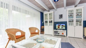 A living room with wicker chairs, a glass coffee table with seashells, a television, and white cabinets with glass doors. - Holiday house beach pearl (Esens)