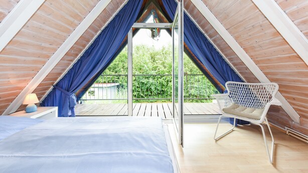 A bedroom with a sloped ceiling, a bed, a chair, and a window overlooking greenery. - Holiday house beach pearl (Esens)