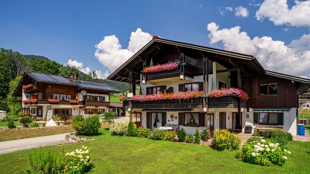 A two-story wooden building with balconies adorned with flowers, surrounded by well-maintained gardens and a clear sky. - Ein-Raum-Wohnung (4) 40 qm, Dusche/WC, Couch, Küchenzeile, Balkon (Reit im Winkl)