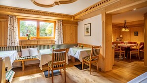 A cozy room with a wooden bench and tables, large windows with curtains, and a dining area with chairs and a table. - Ein-Raum-Wohnung (4) 40 qm, Dusche/WC, Couch, Küchenzeile, Balkon (Reit im Winkl)