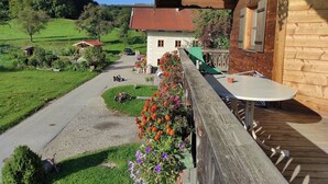 Une terrasse en bois avec une table et des chaises, donnant sur une route de campagne, avec une maison et de la verdure en arrière-plan.