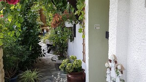 Un camí de pedra estret flanquejat per vegetació exuberant i plantes en testos que condueix a un edifici blanc amb un rètol que diu 'Willkommen'.