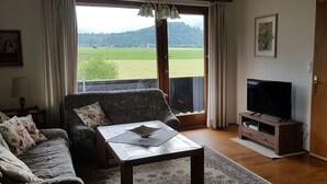 Un saló amb un sofà, una taula de centre, una televisió i vista a un paisatge verd a través de grans finestres.