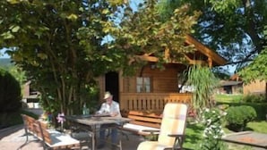 Et udendørs siddeområde med bord og stole, en trækonstruktion med veranda, og en have med en plantekasse.