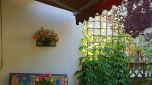A covered patio area with a green table, a colorful chair, a potted plant, and a wall-mounted flower basket. - Quiet vacation apartment, 29sqm, terrace with beautiful garden (Bad Reichenhall)