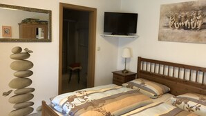 A bedroom with a bed, a TV mounted on the wall, a wooden headboard, a nightstand with a lamp, a mirror, and a painting. - Quiet vacation apartment, 29sqm, terrace with beautiful garden (Bad Reichenhall)