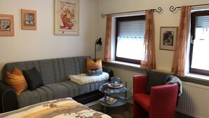 A living room with a grey sofa, red armchair, and a glass coffee table. There are framed pictures on the wall and a window with curtains. - Quiet vacation apartment, 29sqm, terrace with beautiful garden (Bad Reichenhall)